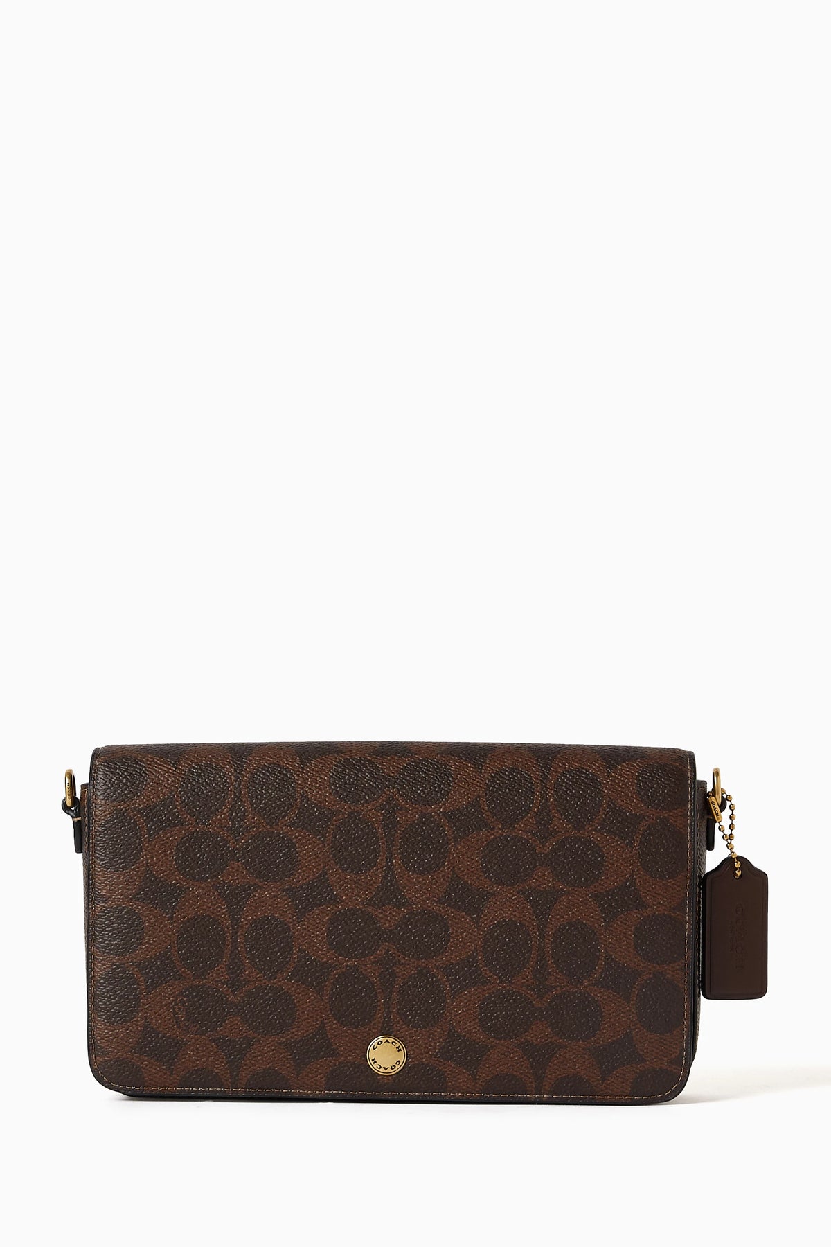 A brown Coach crossbody bag with the signature monogram pattern, featuring a gold-tone front button and a matching leather hangtag on the side, set against a plain white background.
